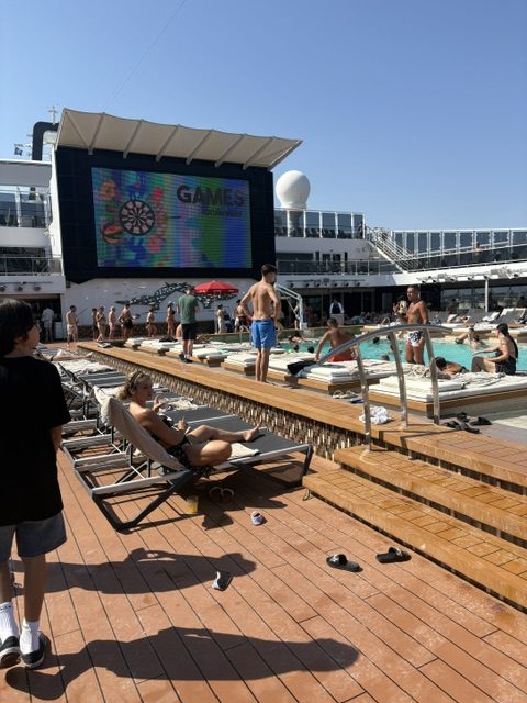 Ship pool deck in bright sun: teak planking, rows of sun loungers, swimmers in the pool, and a large LED screen showing GAMES above the deck