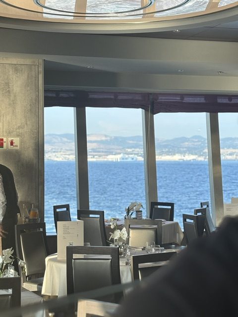 Ship dining room: white-clothed tables with orchid centerpieces and menus, panoramic windows opening onto blue sea and a distant hazy coastline