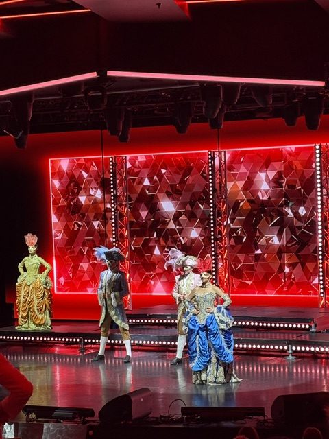 Ship theatre stage lit red, faceted crimson backdrop, performers in baroque costume: a gold gown, a tricorn hat, plumed headdresses and a blue paniered skirt
