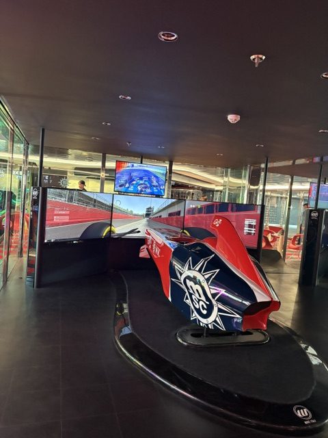 Onboard arcade: a red and blue racing simulator car with the MSC crest on its nose, facing curved track screens in a dark room