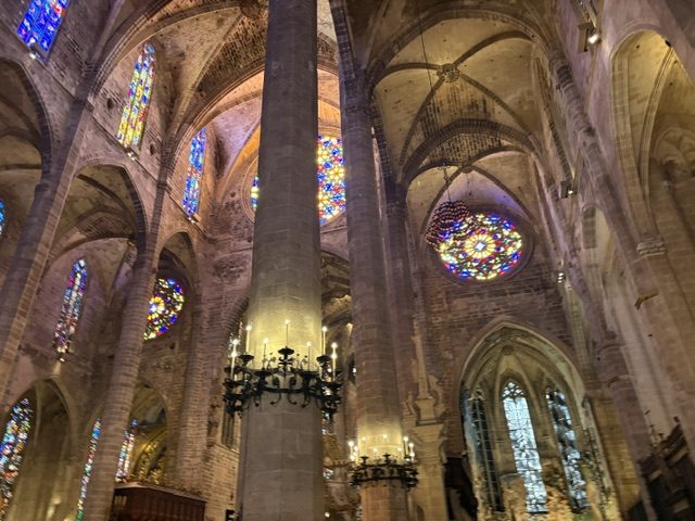 Cathedral interior looking up: a massive stone column, ribbed vaults, stained glass lancets, a rose window and wrought-iron candle chandeliers