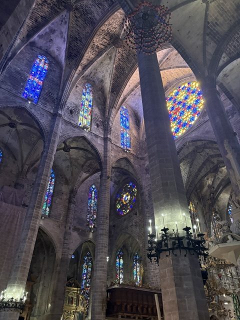 Cathedral nave interior: tall stone columns, deep blue stained glass lancets, two rose windows and iron chandeliers above the choir