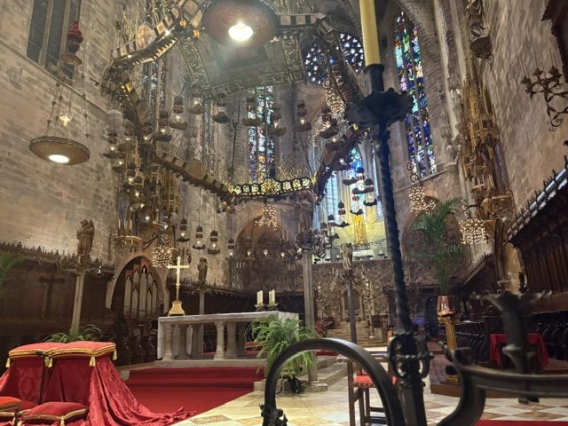 Cathedral sanctuary: an ornate suspended canopy hung with lamps above the altar, red carpet and red-draped seating, carved choir stalls and stained glass behind