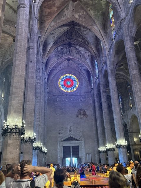 Wide cathedral nave: an enormous multicolored rose window above the west door, towering columns, visitors seated and standing in the foreground