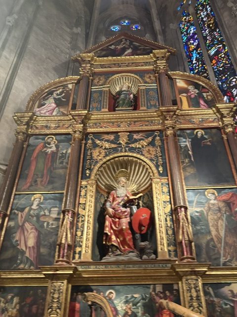 Gilded baroque altarpiece with painted saint panels in tiers and a robed seated figure in a sunburst niche, stained glass window alongside