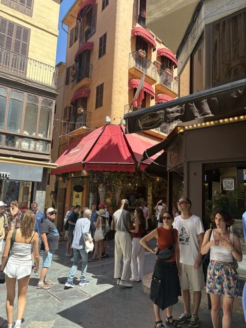 Narrow shopping street in the old center: red awnings over an ornate shopfront, tall shuttered buildings with balconies, crowds of shoppers