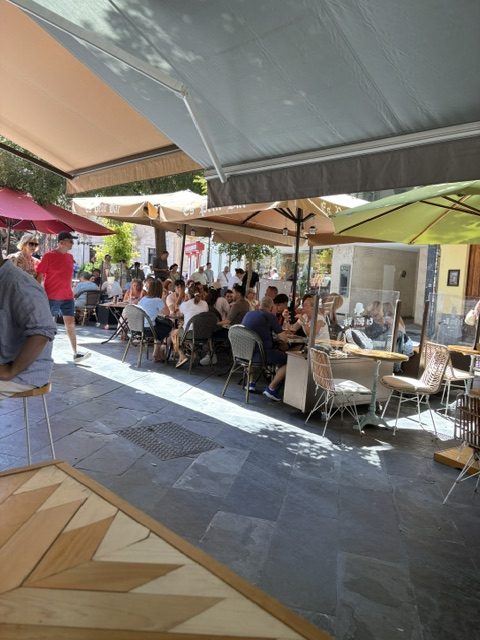 Busy shaded outdoor terrace: peach, red and green awnings and umbrellas over packed tables of diners on grey stone paving