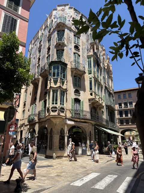 Ornate corner building with a curving tiled facade, green shuttered windows and wrought-iron balconies, pedestrians at the crosswalk below, tree branches framing the sky