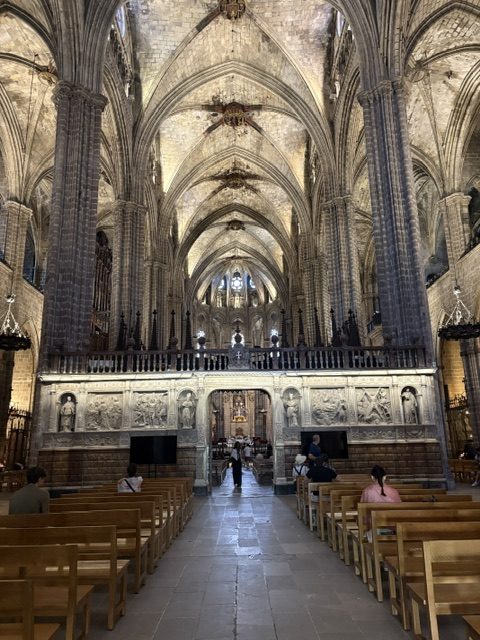 Cathedral nave looking toward the altar: ribbed stone vaults, carved relief choir screen, wooden pews, scattered visitors