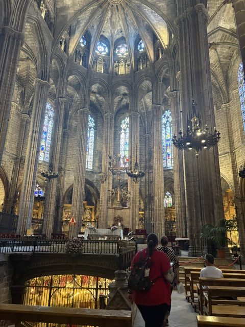 Cathedral apse and ambulatory with tall stained glass windows, iron chandelier, gilded side altars and a lit crypt railing below