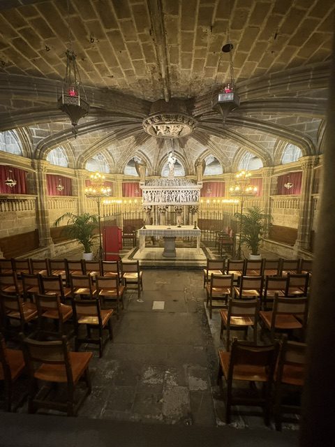Small vaulted chapel with rows of empty wooden chairs facing a candlelit stone altar and red wall hangings