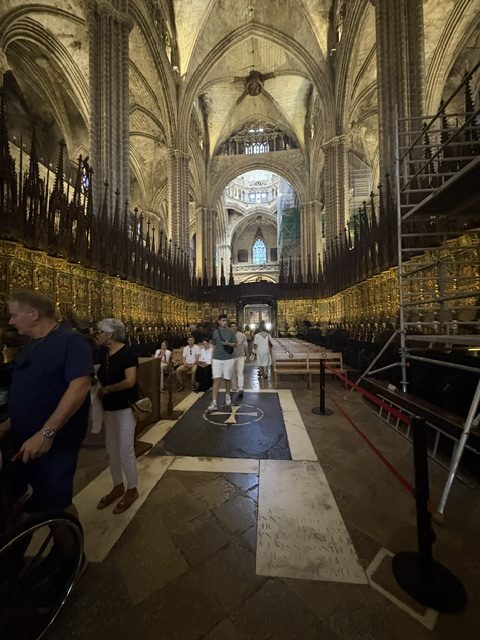 Gilded choir stalls lining the central aisle, restoration scaffolding at right, worn inscribed floor slabs, visitors walking through