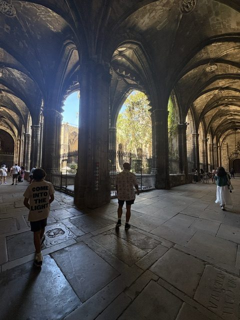 Shadowed cloister arcade with ribbed vaults opening through iron grilles onto a sunlit green courtyard; two people walking toward the light