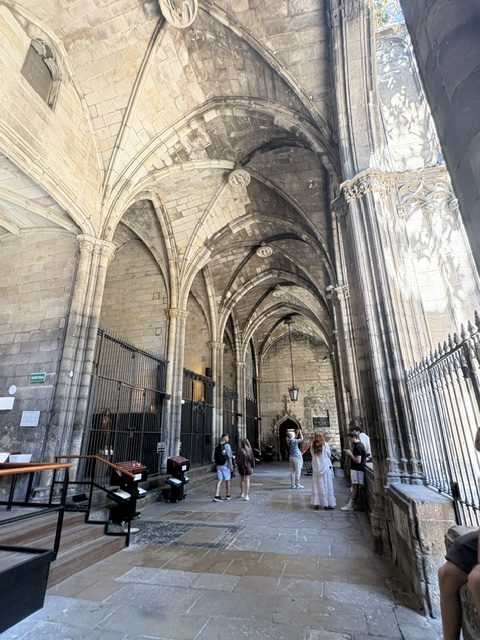Long vaulted cloister gallery with stone columns, iron grilles, a hanging lantern and visitors along the flagstones
