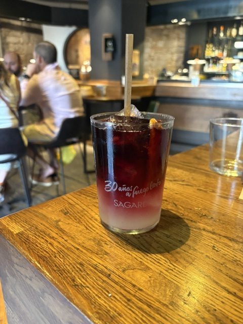 Tall glass of dark red sangria with fruit and a wooden stirrer on a wooden bar top, Sagardi lettering on the glass, diners behind
