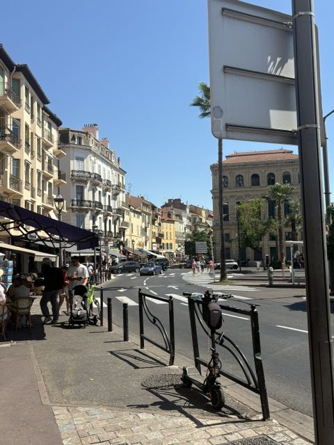 Street corner with cafe awnings and outdoor tables, an electric scooter at a rack, apartment buildings lining the road