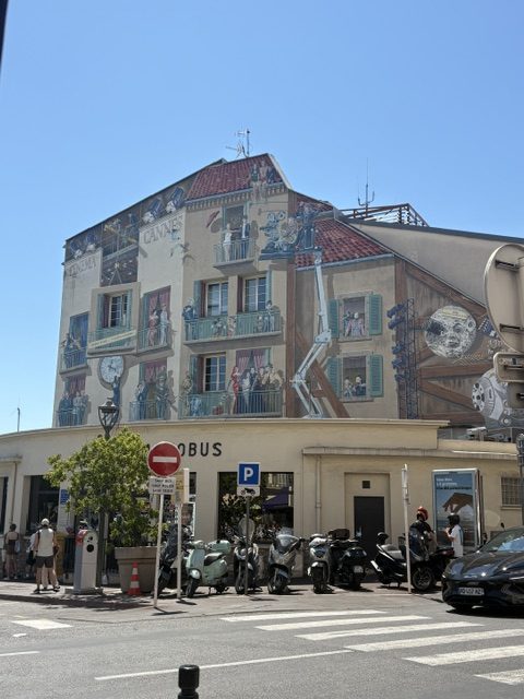 Building facade painted with a large trompe-l'oeil mural of figures on balconies above a bus stop, motorbikes and scooters parked below