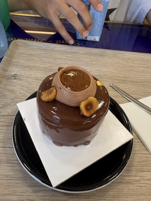 Glossy chocolate mousse cake topped with a small clay pot of caramel and hazelnut discs, on a black plate with a fork