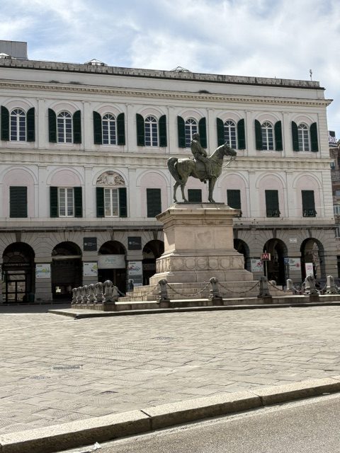 Bronze equestrian statue on a tall stone plinth in a piazza, shuttered palazzo with arcade behind