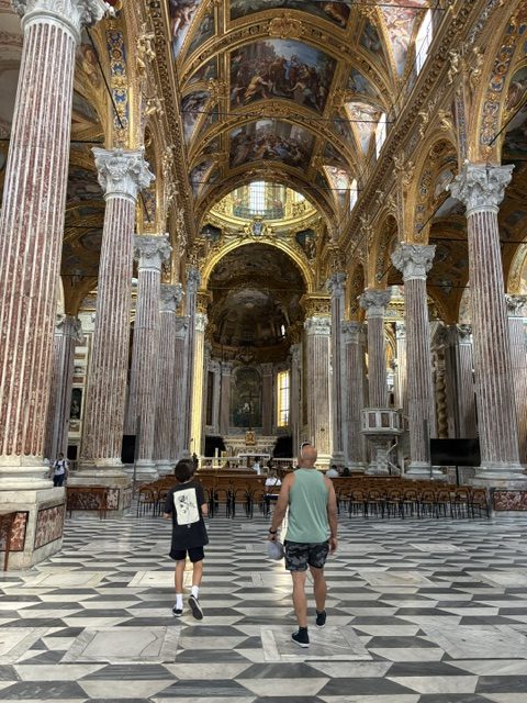 Church nave with tall marble columns and checkerboard marble floor, two visitors walking toward the altar