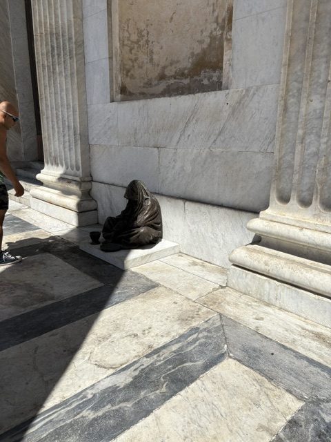 Bronze sculpture of a shrouded seated beggar with a bowl, on a marble church portico floor, passerby at left