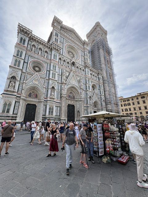 Duomo facade in green, pink and white marble, dense crowd and souvenir stalls in the piazza