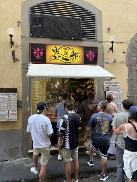Queue of people on the street outside the I' Girone De' Ghiotti sandwich shop under its yellow sign