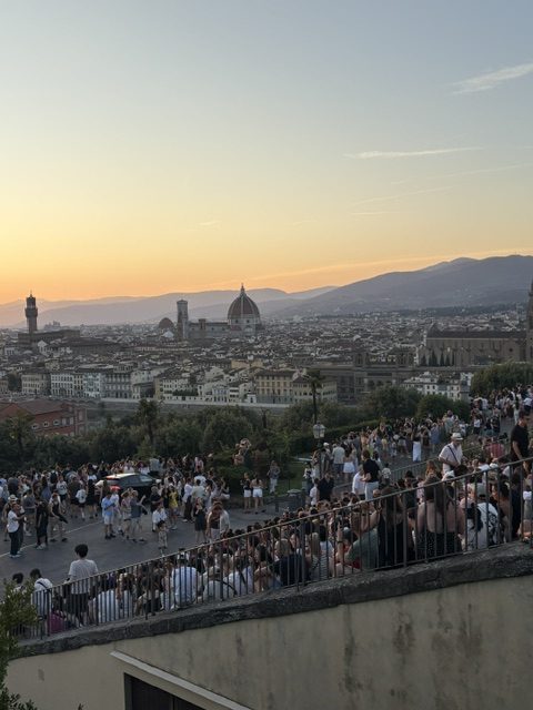 Wide sunset view over Florence from a crowded hilltop terrace, people lining the wall below