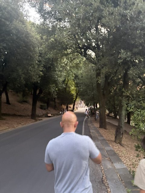 Man in a white shirt walking down a shaded tree-lined road descending toward the city