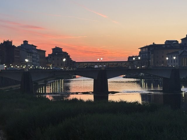 Sunset over the Arno with bridges silhouetted, pink and orange sky, dark riverbank foreground