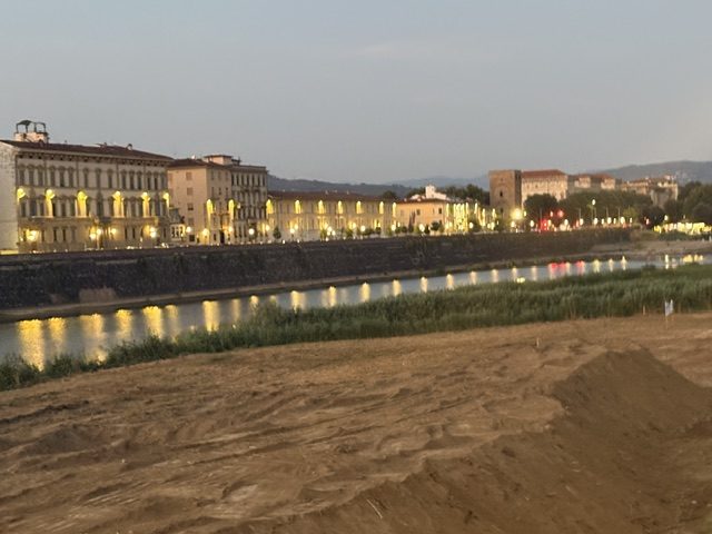 Riverside buildings lit at dusk along the Arno, gravel bank in the foreground