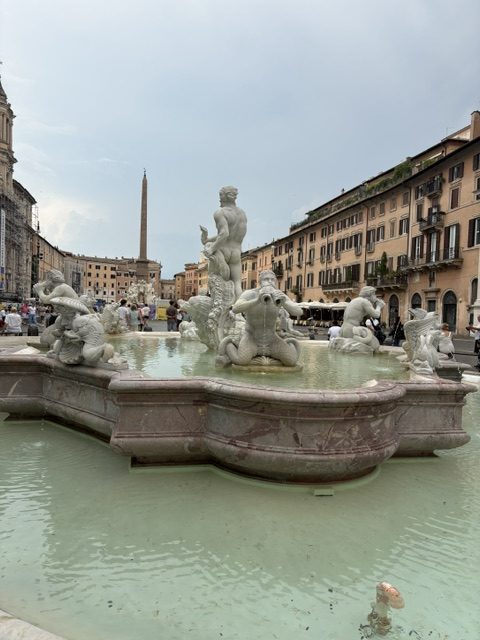Baroque fountain with marble sea gods and a triton, pale green basin water, obelisk and palazzi behind, Piazza Navona