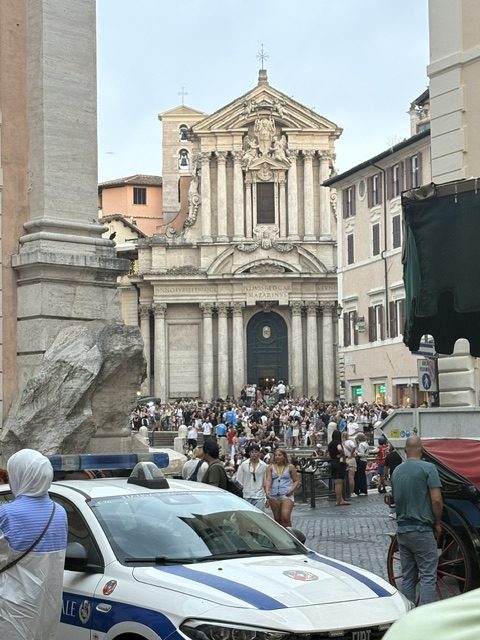 Baroque church facade with columns and bell tower, packed crowd on the steps below, blue and white police car and a horse carriage in the foreground