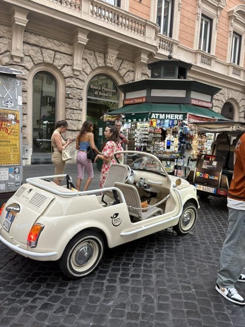 Cream vintage Fiat 500 open-top with wicker seats parked on cobbles beside a green newsstand kiosk
