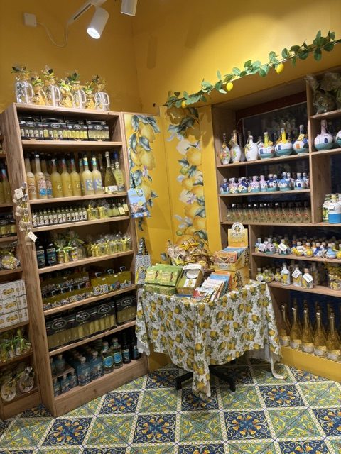 Shop interior with yellow walls, wooden shelves of limoncello and lemon products, lemon garlands, lemon-print tablecloth, patterned majolica floor tile