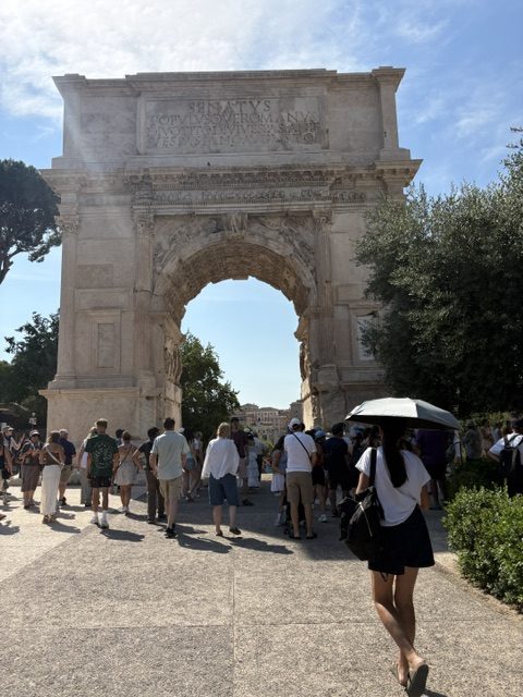 Single-bay Roman triumphal arch against blue sky, visitors below, one walking under a black sun umbrella