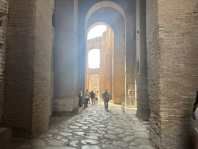 Shadowed brick arcade passage with massive piers, ancient cobbled paving, sunlit brick ruins visible through the arch ahead