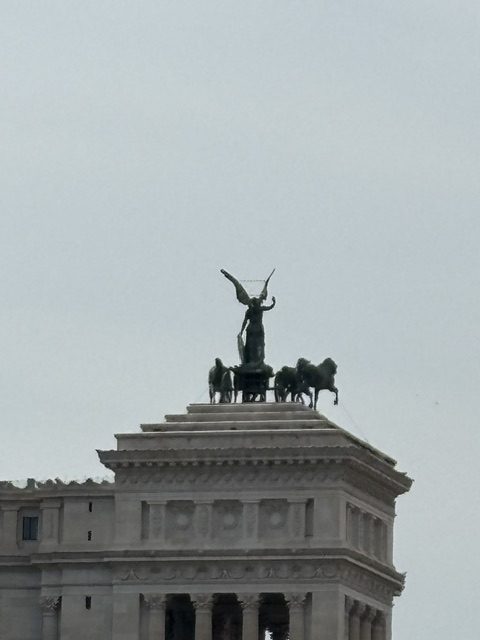 Bronze winged Victory driving a chariot with horses on the roofline of a white marble monument, grey sky