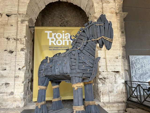 Slatted wooden horse sculpture standing in a stone niche beside a yellow exhibition banner reading Troia Roma