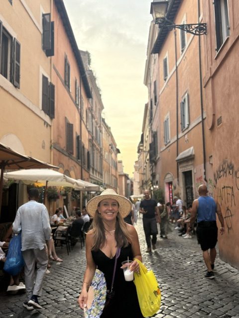 Woman in a straw hat and black dress smiling in a narrow Trastevere lane at golden hour, holding a cold drink and a yellow shopping bag, cafe tables under umbrellas