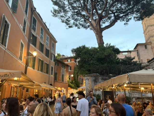 Evening piazza with a large umbrella pine, rows of awnings and outdoor tables full of diners, church wall at right