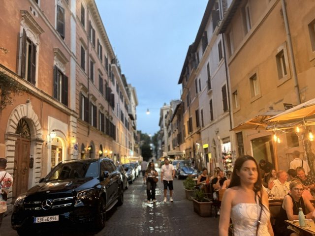 Narrow Trastevere street at dusk, parked cars, lit cafe terraces on both sides, people walking the center